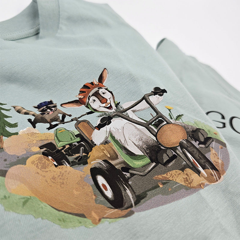 Nahaufnahme des T-Shirt-Motivs auf grüner Vorderseite: Ziege und Waschbär flitzen gemeinsam im Go-Kart, farbenfroh und kindgerecht.