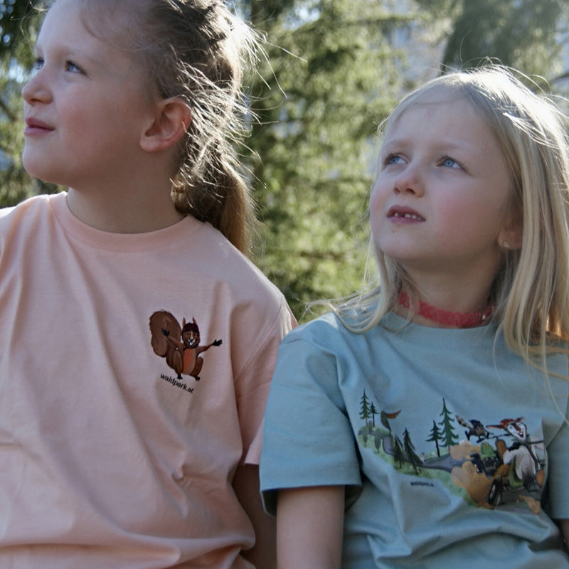 Kind im rosa Go Wild T-Shirt im Wald, Vorderseite sichtbar und zweites Kind im grünen Go Wild T-Shirt robustes Bio-Baumwoll-Shirt für Abenteuer und Ausflüge