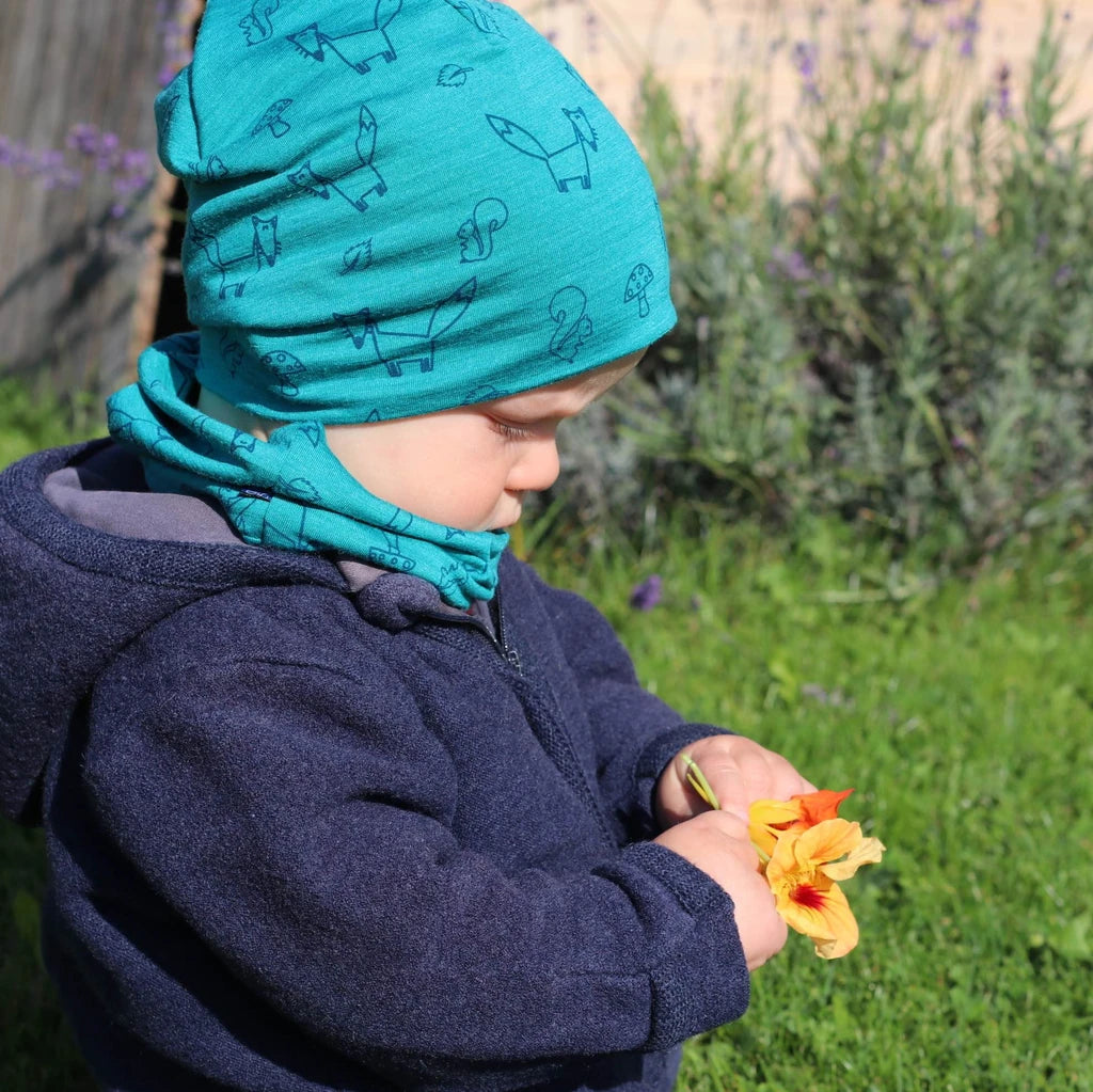Kind trägt smaragdfarbene Merino-Seide-Haube mit Fuchs-Motiv im Garten, kuschelig weiche und temperaturausgleichende Kinderhaube für draußen