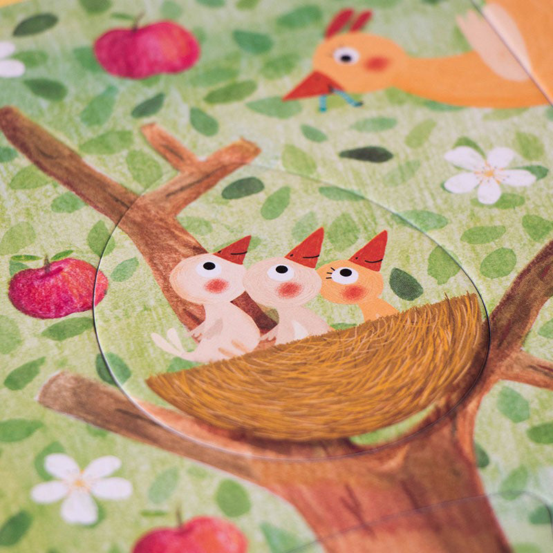 Detailansicht eines Puzzleteils mit drei kleinen Vögeln im Nest, darüber ein gelber Vogel mit rotem Schnabel, grüne Blätter und Äpfel im Hintergrund.