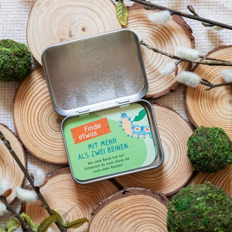 Geöffnete Metallbox mit einer Karte, auf der steht „Finde etwas mit mehr als zwei Beinen“ – illustriert mit einer Raupe.