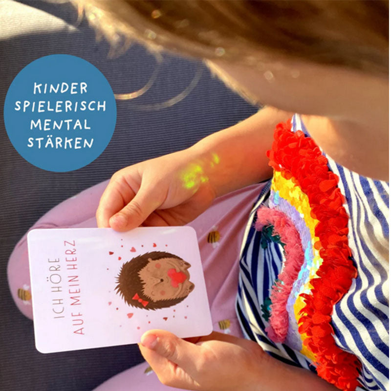 Kind hält eine Affirmationskarte mit Igelillustration und der Botschaft Ich höre auf mein Herz, farbenfrohe Kleidung und weiches Licht im Alltagsszenario