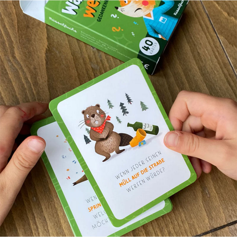 Nahaufnahme von Kinderhänden, die mehrere Fragekarten fächern, oben eine Karte mit Bärenillustration und Frage zum Thema Umwelt, Kartenbox im Hintergrund auf Holztisch