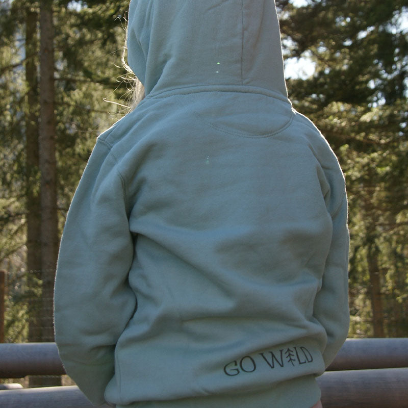 Rückansicht eines Kindes im Wald, mit aufgesetzter Kapuze. Kind trägt hell türkisgrünen Bio-Baumwollhoody, gemütliche und atmungsaktive Kleidung für kleine Entdecker