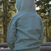 Rückansicht eines Kindes im Wald, mit aufgesetzter Kapuze. Kind trägt hell türkisgrünen Bio-Baumwollhoody, gemütliche und atmungsaktive Kleidung für kleine Entdecker