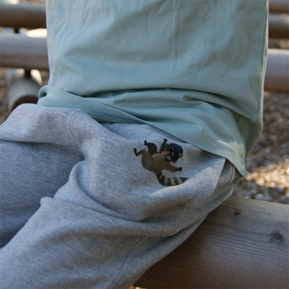 Nahaufnahme der grauen Bio-Baumwollhose mit Waschbären-Print, zeigt weichen Stoff und sorgfältige Verarbeitung, getragen von einem Kind im Grünen