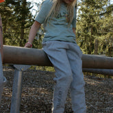 Kind trägt graue Bio-Baumwollhose mit Waschbären-Print und Sturzhelm beim Spielen im Wald, bequeme und nachhaltige Kinderkleidung für draußen.