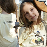 Kind trägt gelbes Go Wild T-Shirt im Wald, Vorderseite mit Waschbär-Motiv im Go-Kart, aktiv beim Spielen und Entdecken