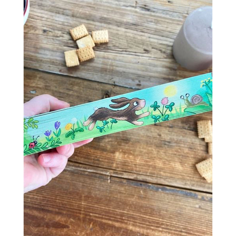 Hand hält ein bunt illustriertes Holzstück mit Hase, Blumen, Klee und Schnecke, im Hintergrund ein Holztisch mit Snacks und Kerze.
