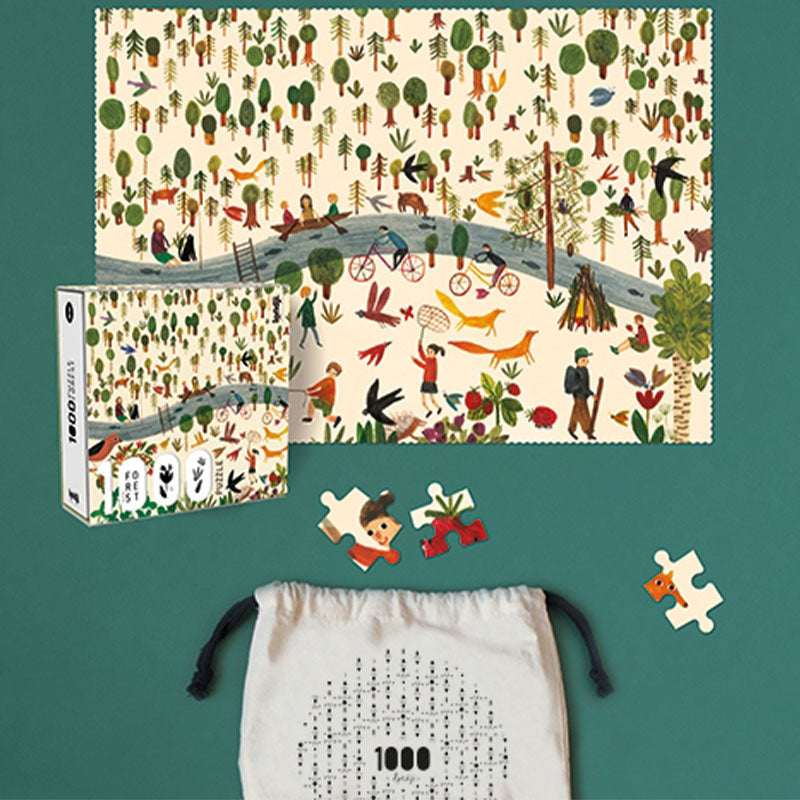 Fertig zusammengesetztes 1000-Teile-Puzzle mit Waldmotiv: zahlreiche grüne und braune Bäume, Menschen beim Paddeln im Kanu, Radfahrer auf einem Weg, Spaziergänger, spielende Kinder, ein Fuchs, Vögel und verschiedene Tiere; daneben die Puzzlebox und ein weißer Stoffbeutel mit schwarzem Aufdruck, aus dem Puzzleteile herausragen.