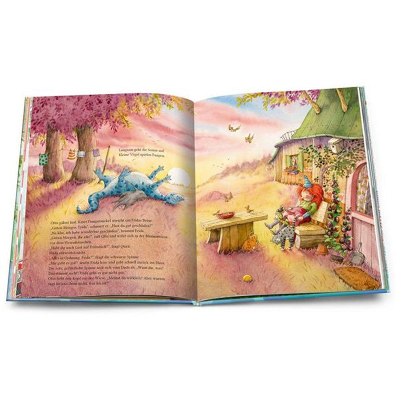 Buchseite – Frida sitzt draußen vor ihrem kleinen Hexenhaus an einem Holztisch, neben ihr der blaue Drache und ihre Katze, bunte Herbstlandschaft im Hintergrund.