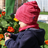 Kind trägt himbeerfarbene Merino-Seide-Haube mit Geißlein-Motiv beim Spielen im Garten, warme und nachhaltige Kinderkleidung in der Natur.