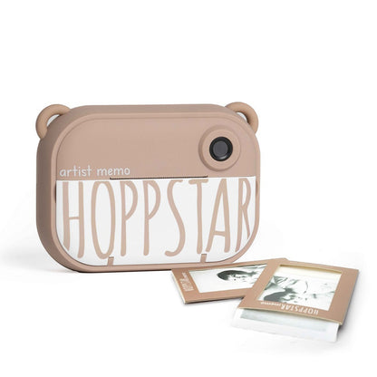 Hoppstar Artist Memo Kamera mit zwei ausgedruckten Schwarz-Weiß-Fotos davor auf weißem Hintergrund.