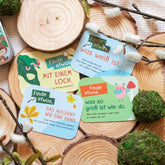 Mehrere bunte Spielkarten mit Suchaufgaben wie „Finde etwas mit einem Loch“ und „Finde etwas, das aussieht wie eine Hand“, dekorativ auf Holzscheiben arrangiert.