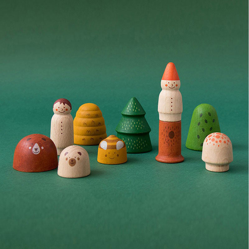 Mehrere bemalte Holzfiguren aus dem Set „My Wooden World – Forest“ auf grünem Hintergrund, darunter Bär, Kind, Pilze, Bäume, Bienenstock und Zwerg.