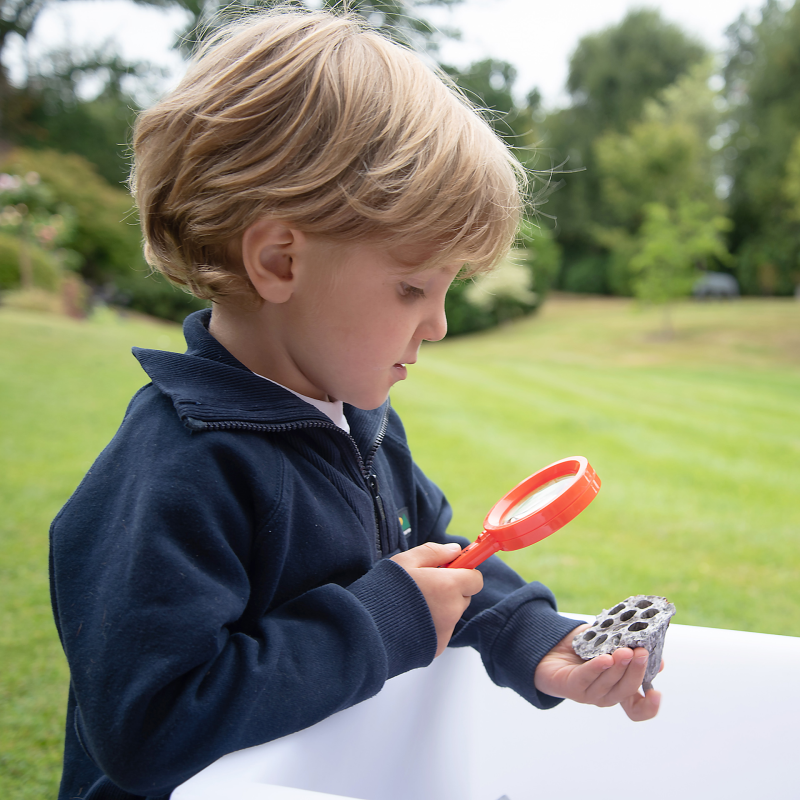 Kind sitzt draußen auf der Wiese und schaut durch die Regenbogenlupe durch