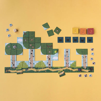 Spielaufbau auf gelbem Hintergrund mit Karten, Würfeln und Spielfiguren; zeigt den Wald mit Bäumen und Wegen.