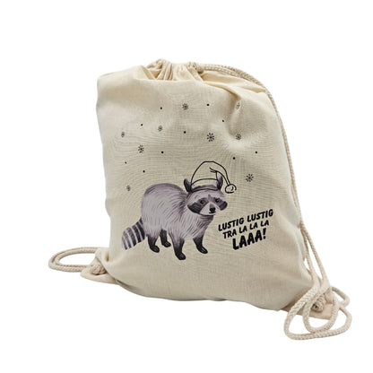 Beiger Stoffturnbeutel mit Kordelzug, bedruckt mit einem Waschbären mit Weihnachtsmütze und Schneeflocken. Daneben der Schriftzug „Lustig lustig tra la la la laaa!“.