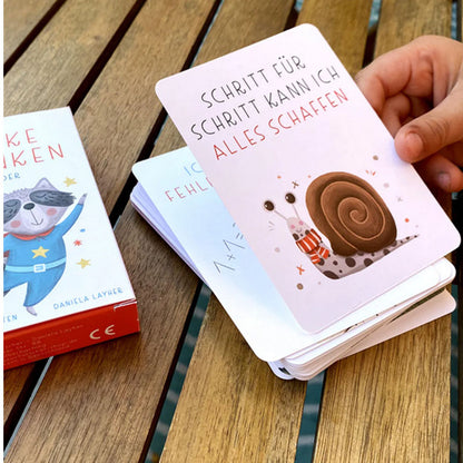 Hand hält eine Affirmationskarte mit Schneckenillustration und dem Text Schritt für Schritt kann ich alles schaffen, Kartenbox und weitere Karten auf Holztisch im Hintergrund