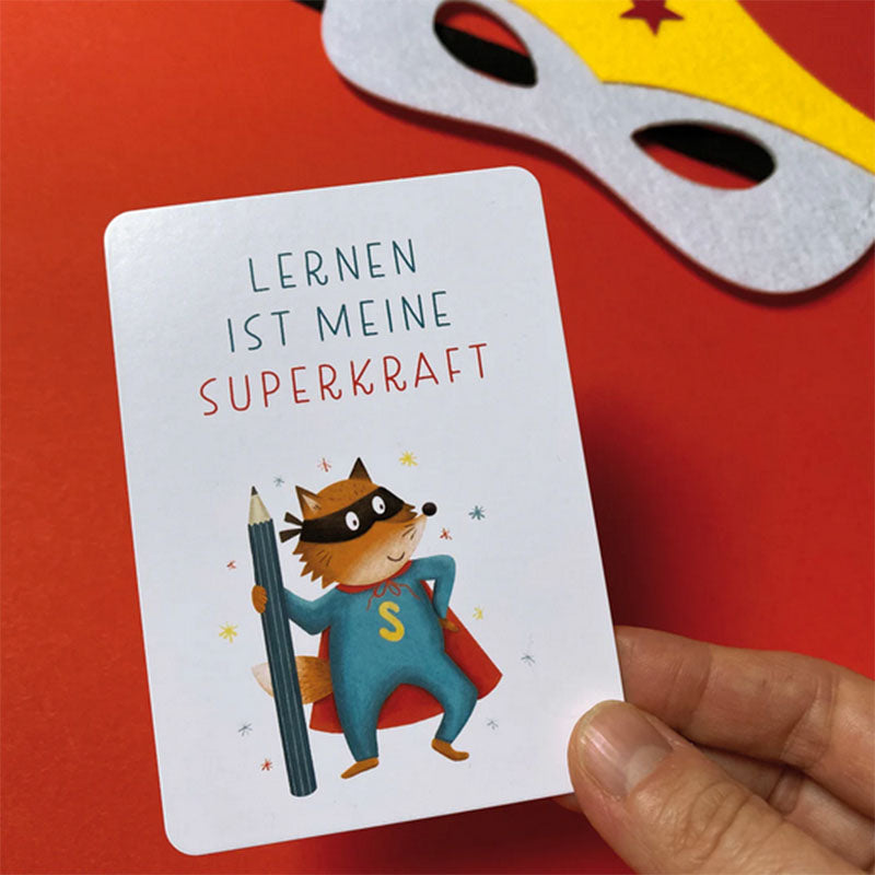 Nahaufnahme einer einzelnen Affirmationskarte in der Hand mit Illustration eines Superhelden-Tiers mit Stift und dem Text Lernen ist meine Superkraft auf rotem Hintergrund