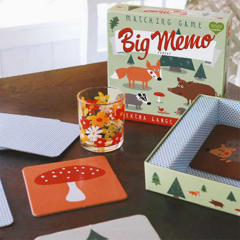 Spielsituation: geöffnete Spielschachtel, ausgelegte Karten mit Waldmotiven und ein Glas mit Blumenmuster auf einem Tisch.