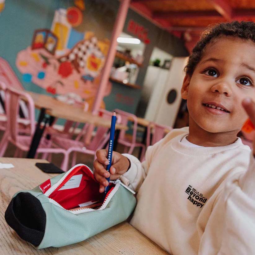 Kind holt blauen Stift aus der Federmappe in der Farbe Mini Pistache die am Tisch liegt