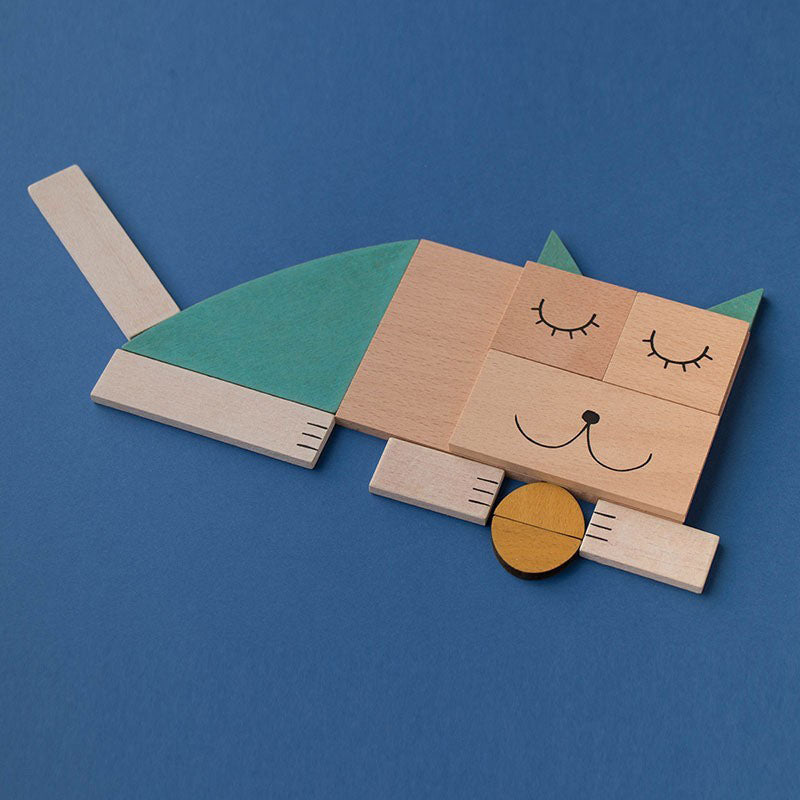 Schlafende Katze aus Holzformen mit grünem Rücken und gelber Kugel vor der Schnauze auf blauem Hintergrund.