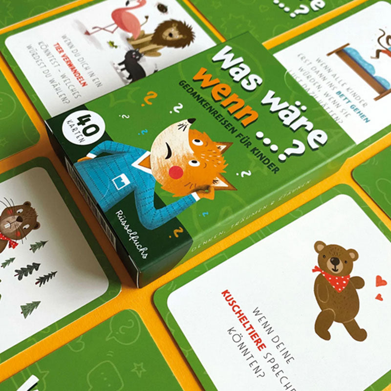 Schräg liegende Kartenbox Was wäre wenn umgeben von mehreren offenen Fragekarten mit Tierillustrationen und kurzen Fragen, angeordnet auf gelb-grünem Untergrund