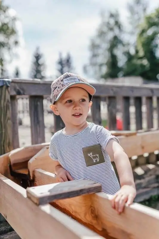 Junge mit Kapperl steht im Wasserspielplatz an einem Wasserlauf aus Holz und beobachtet das Wasser beim Fließen