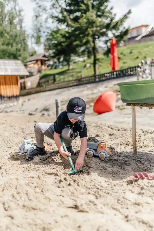 Junge mit Kapperl am Boden auf dem Sand am Wasserspielplatz auf der Teichalm beim Rechen und Befüllen eines kleinen Baggers mit Sand