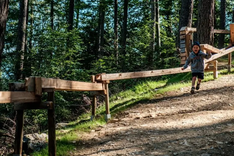 Ein Junge mit Kapperl und kurzer Hose läuft mit offenem Mund im Schatten des Waldes neben der Waldkugelbahn her.